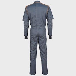 May include: A slate gray jumpsuit featuring a high collar, brown shoulder accents, and long sleeves with black ribbed cuffs. The forearms display a white geometric triangle pattern, while the back includes vertical seam detailing and a central closure.