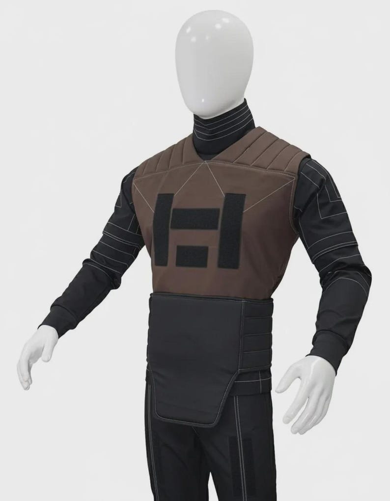 May include: A brown and black tactical vest worn over a black long-sleeve shirt with white geometric line detailing. The vest features a quilted upper chest panel, a central black rectangular emblem, and a solid black lower abdominal guard panel.