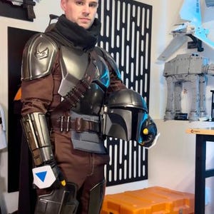 May include: A full-body costume featuring metallic silver shoulder and leg armor, a brown jumpsuit, a black neck gaiter, and a dark grey helmet with a T-shaped visor. The armor includes a chest plate, gauntlets, and a belt with ammunition pouches.