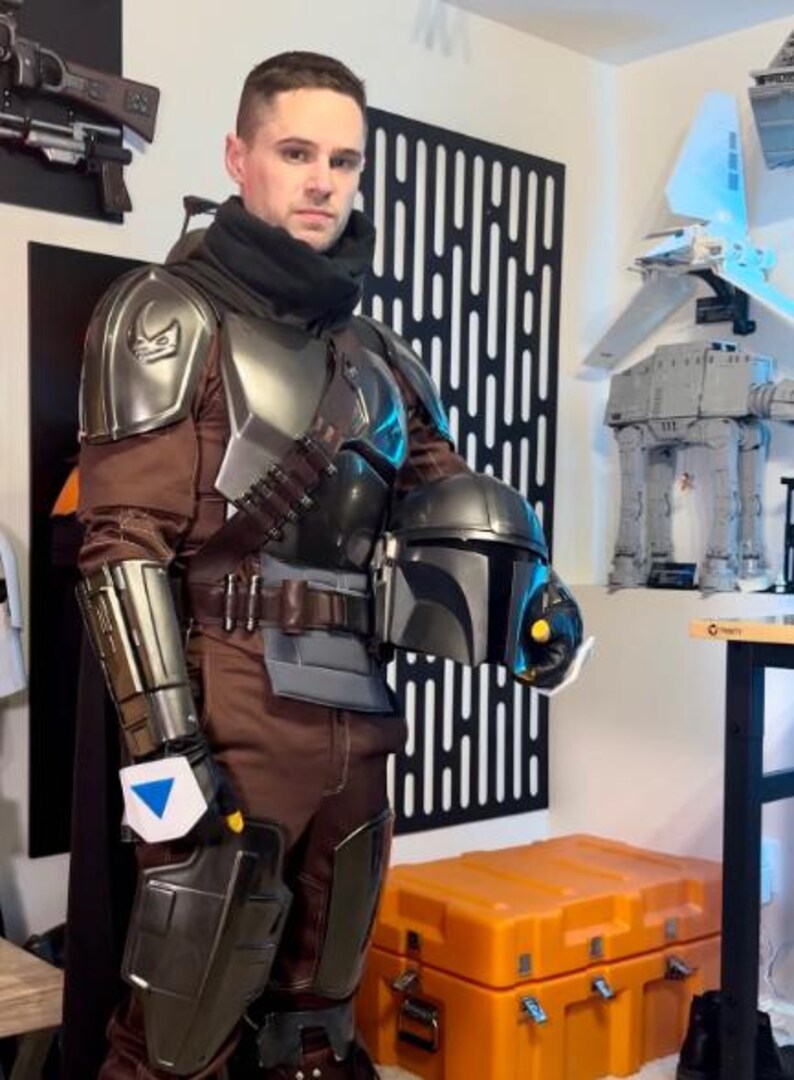 May include: A full-body costume featuring metallic silver shoulder and leg armor, a brown jumpsuit, a black neck gaiter, and a dark grey helmet with a T-shaped visor. The armor includes a chest plate, gauntlets, and a belt with ammunition pouches.