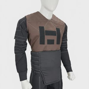 May include: A brown chest plate with black rectangular patches worn over a black long-sleeved shirt featuring white contrast stitching. The garment includes a black padded waist belt and black trousers with matching white stitching details.