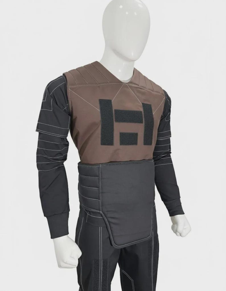 May include: A brown chest plate with black rectangular patches worn over a black long-sleeved shirt featuring white contrast stitching. The garment includes a black padded waist belt and black trousers with matching white stitching details.