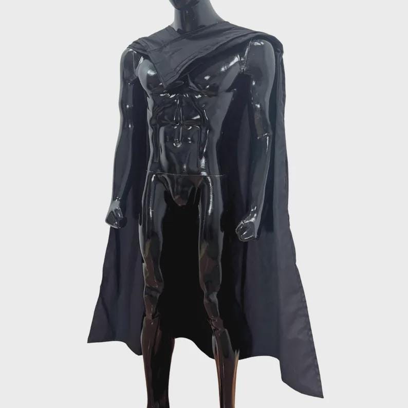 Puede incluir: Una capa negra drapeada sobre un maniqu&iacute; negro brillante. La capa presenta un cuello estructurado que descansa sobre los hombros y se extiende por la espalda, cubriendo el torso y las piernas con un acabado de tela negra lisa y s&oacute;lida.