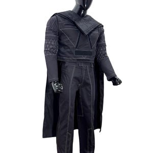 May include: A black long-sleeved top and matching trousers set featuring white contrast stitching and a geometric grid pattern on the sleeves. The outfit includes a draped shoulder piece and a cape, all displayed on a black mannequin torso.