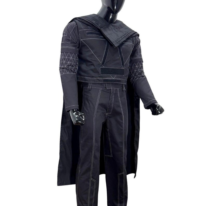 May include: A black long-sleeved top and matching trousers set featuring white contrast stitching and a geometric grid pattern on the sleeves. The outfit includes a draped shoulder piece and a cape, all displayed on a black mannequin torso.
