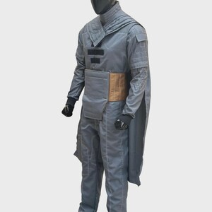 Puede incluir: Un mono gris de manga larga con cuello alto, un panel de cintura color canela y una capa unida a los hombros. La prenda incluye detalles rectangulares negros en el pecho, costuras blancas visibles y guantes negros en el maniqu&iacute;.