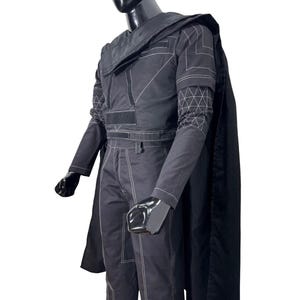 May include: A black long-sleeved jacket and matching trousers set featuring white contrast stitching, a structured collar, and a long cape. The jacket includes geometric white line patterns on the sleeves and rectangular fabric panels on the chest.