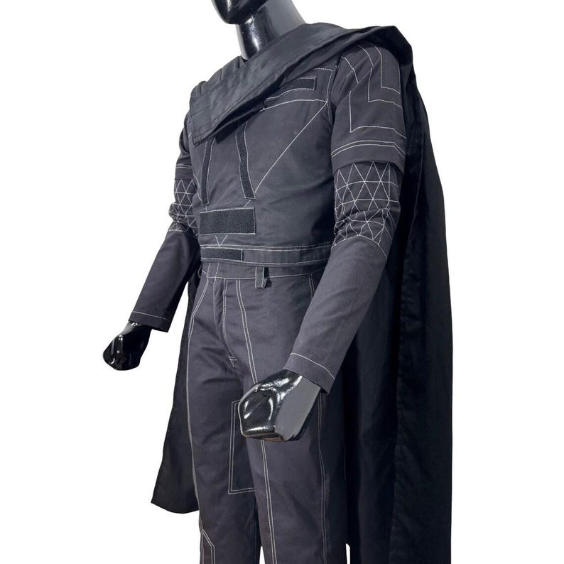 May include: A black long-sleeved jacket and matching trousers set featuring white contrast stitching, a structured collar, and a long cape. The jacket includes geometric white line patterns on the sleeves and rectangular fabric panels on the chest.