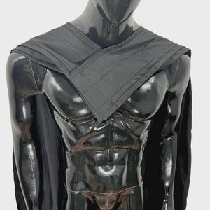 Puede incluir: Una capa negra con un cuello plisado y estructurado que se cruza sobre el pecho. La prenda est&aacute; colocada sobre los hombros de un maniqu&iacute; negro brillante, mostrando una tela negra s&oacute;lida con l&iacute;neas rectas y un dise&ntilde;o sencillo.