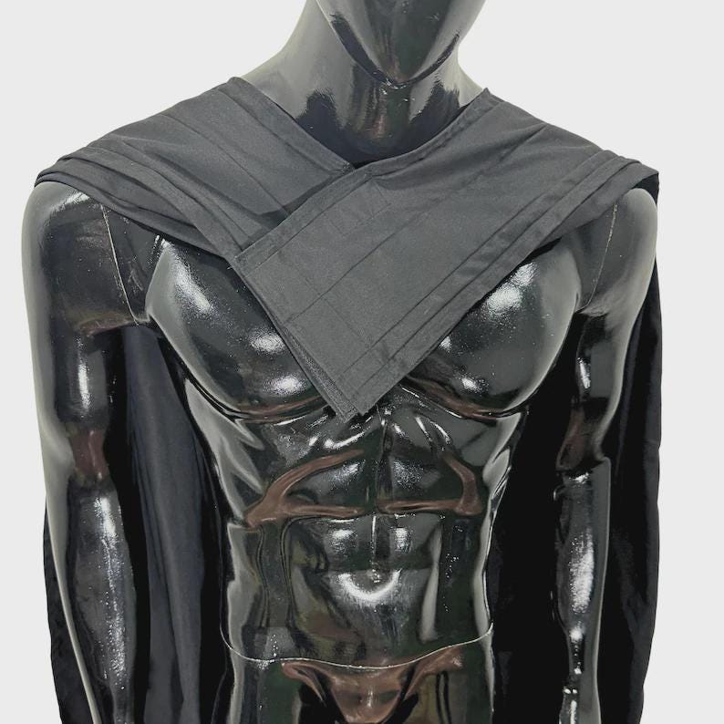 Puede incluir: Una capa negra con un cuello plisado y estructurado que se cruza sobre el pecho. La prenda est&aacute; colocada sobre los hombros de un maniqu&iacute; negro brillante, mostrando una tela negra s&oacute;lida con l&iacute;neas rectas y un dise&ntilde;o sencillo.