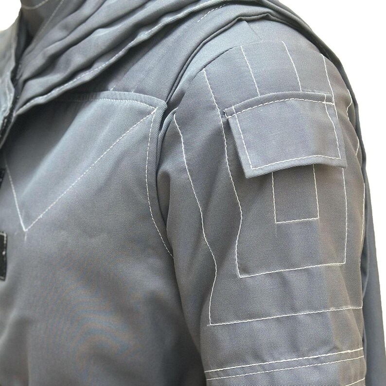 Puede incluir: Una chaqueta gris con costuras blancas prominentes que delinean los bordes y un bolsillo rectangular en la parte superior de la manga. La prenda incluye un cuello estructurado y un dise&ntilde;o de panel en forma de V en el pecho con costuras.