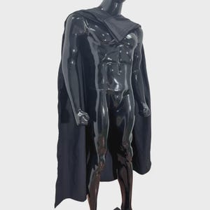 Puede incluir: Una capa negra drapeada sobre un maniqu&iacute; negro brillante. La capa presenta una pieza de cuello estructurada que descansa sobre los hombros y la parte superior del pecho, extendi&eacute;ndose por la espalda y los lados en un dise&ntilde;o de tela negra s&oacute;lida y fluida.