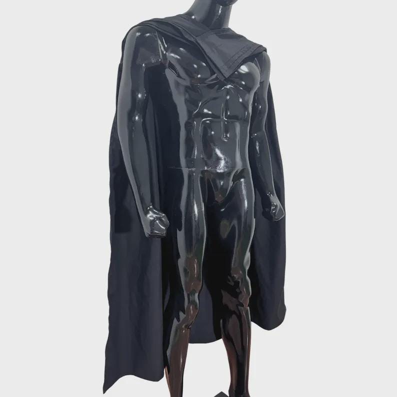 Puede incluir: Una capa negra drapeada sobre un maniqu&iacute; negro brillante. La capa presenta una pieza de cuello estructurada que descansa sobre los hombros y la parte superior del pecho, extendi&eacute;ndose por la espalda y los lados en un dise&ntilde;o de tela negra s&oacute;lida y fluida.