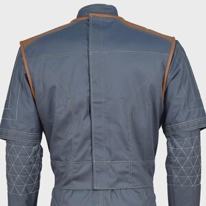 May include: A slate blue jacket featuring brown leather-like trim along the shoulders and upper back. The garment includes a high collar, visible white topstitching, and a geometric triangular quilted pattern on the lower sleeves and cuffs.