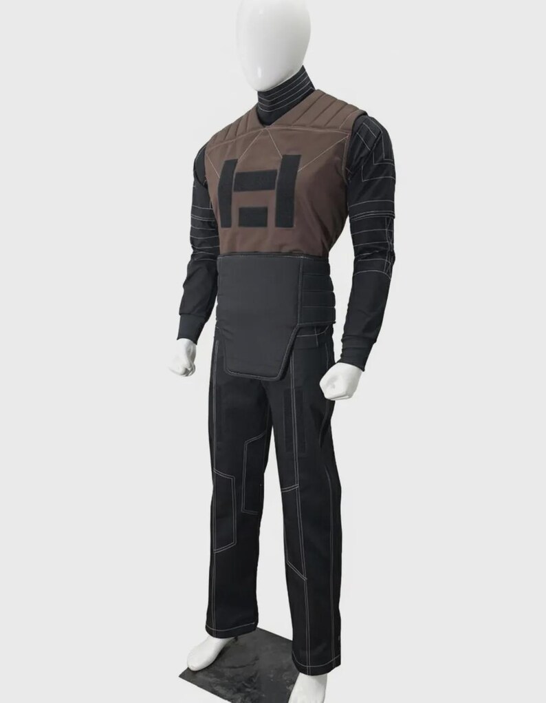 May include: A costume set on a mannequin featuring a brown quilted vest with a black geometric emblem, worn over a black long-sleeve shirt with white line patterns. The outfit includes a black waist guard and matching black trousers with white stitching.