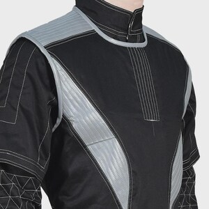 Puede incluir: Una chaqueta negra de manga larga con cuello alto y paneles acolchados de color gris plateado en los hombros y el pecho. La prenda incluye costuras blancas en contraste y un patr&oacute;n geom&eacute;trico de tri&aacute;ngulos en la parte inferior de las mangas.