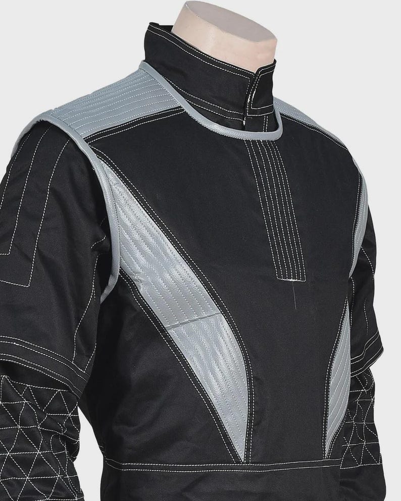 Puede incluir: Una chaqueta negra de manga larga con cuello alto y paneles acolchados de color gris plateado en los hombros y el pecho. La prenda incluye costuras blancas en contraste y un patr&oacute;n geom&eacute;trico de tri&aacute;ngulos en la parte inferior de las mangas.