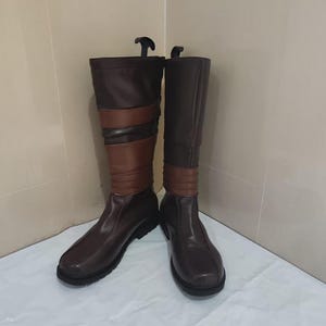 May include: A pair of knee-high boots in dark brown with a lighter brown horizontal band detail around the mid-calf. The boots feature a rounded toe, a flat black sole, and a vertical seam running down the center of the foot and shaft area.