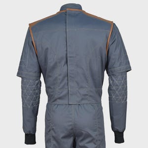 May include: A slate blue jacket with brown leather-like shoulder accents and white contrast stitching. The sleeves feature a quilted triangular pattern on the forearms, a high collar, and black ribbed cuffs. The back displays a vertical center seam.