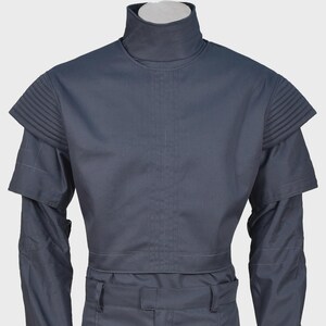 Peut inclure: Une tunique gris fonc&eacute; &agrave; col montant dot&eacute;e d'&eacute;paulettes pliss&eacute;es structur&eacute;es et de manches longues. Le v&ecirc;tement pr&eacute;sente un d&eacute;tail de couture verticale centrale sur la poitrine et une ceinture assortie avec passants, offrant un aspect uniforme.