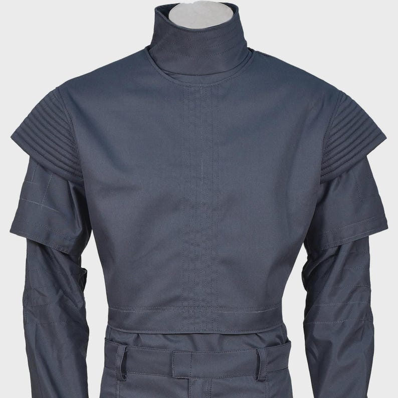 Peut inclure: Une tunique gris fonc&eacute; &agrave; col montant dot&eacute;e d'&eacute;paulettes pliss&eacute;es structur&eacute;es et de manches longues. Le v&ecirc;tement pr&eacute;sente un d&eacute;tail de couture verticale centrale sur la poitrine et une ceinture assortie avec passants, offrant un aspect uniforme.