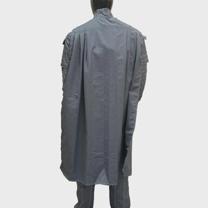 Puede incluir: Una capa larga de color gris sobre una chaqueta gris a juego con detalles acolchados en los hombros. La prenda presenta un cuello alto y un dise&ntilde;o estructurado y fluido que cae por la espalda, cubriendo completamente los hombros y el torso.
