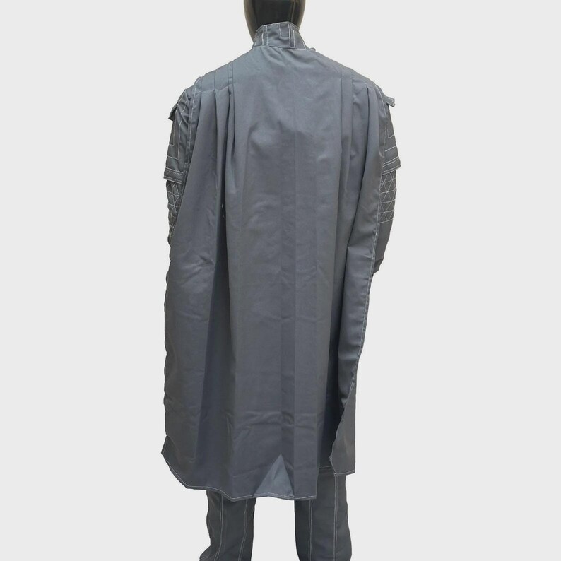 Puede incluir: Una capa larga de color gris sobre una chaqueta gris a juego con detalles acolchados en los hombros. La prenda presenta un cuello alto y un dise&ntilde;o estructurado y fluido que cae por la espalda, cubriendo completamente los hombros y el torso.