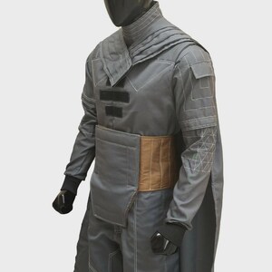 Puede incluir: Una t&uacute;nica gris de manga larga con cuello alto, con patrones geom&eacute;tricos cosidos en blanco en las mangas y hombros, detalles rectangulares negros en el pecho y un panel de cintura acolchado de color tostado sobre pantalones a juego.