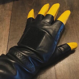 May include: A black leather glove featuring bright yellow fingertips and a thumb tip. The glove includes a rectangular black hook-and-loop fastener patch on the back of the hand and a gathered, elasticized wrist section with visible stitching.