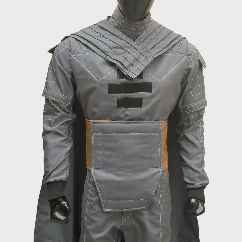 Puede incluir: Una chaqueta de disfraz gris de manga larga con cuello alto, paneles de hombro plisados y un panel de cintura rectangular de color tostado. La prenda incluye detalles rectangulares negros en el pecho, costuras blancas y pu&ntilde;os negros.