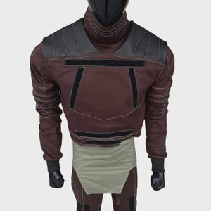 Puede incluir: Una chaqueta marr&oacute;n de manga larga con cuello alto, paneles negros en los hombros y tiras de velcro horizontales negras en el pecho. La chaqueta presenta costuras blancas en contraste y un panel de cintura en color verde oliva claro.