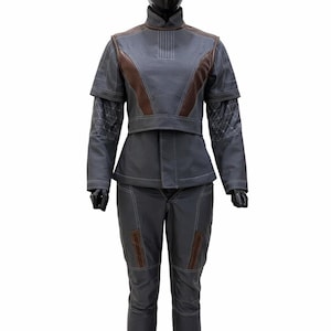 May include: A dark grey two-piece outfit featuring a high-neck jacket with brown accents, white contrast stitching, and geometric patterns on the sleeves. The matching trousers include brown vertical panels and white stitching details throughout.