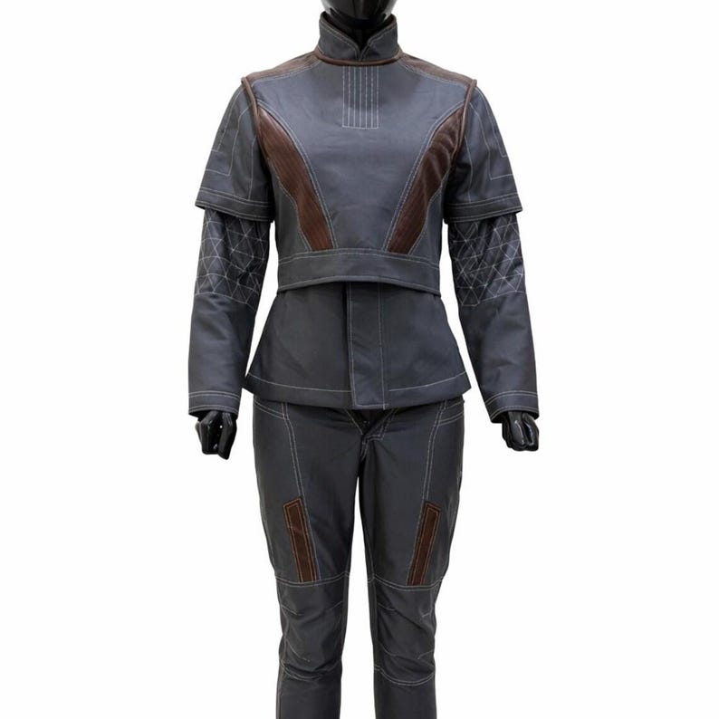 May include: A dark grey two-piece outfit featuring a high-neck jacket with brown accents, white contrast stitching, and geometric patterns on the sleeves. The matching trousers include brown vertical panels and white stitching details throughout.