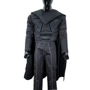 May include: A black costume set featuring a long-sleeved top with white geometric stitching, a matching pair of trousers with white seam detailing, and a long black cape. The top includes rectangular velcro patches on the chest and shoulders.
