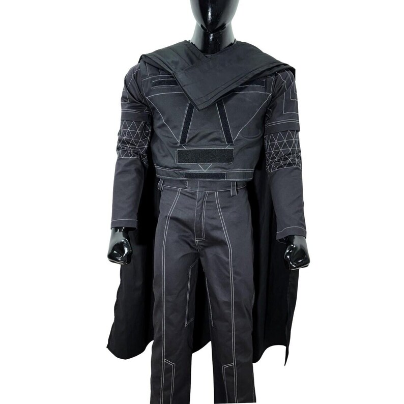 May include: A black costume set featuring a long-sleeved top with white geometric stitching, a matching pair of trousers with white seam detailing, and a long black cape. The top includes rectangular velcro patches on the chest and shoulders.