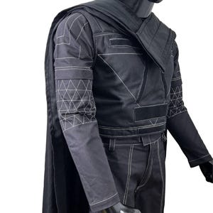 May include: A black long-sleeved jacket and matching trousers set featuring white contrast stitching. The jacket includes a structured collar, a cape attached at the shoulders, and geometric triangle patterns on the sleeves. Black velcro strips are visible on the chest area.