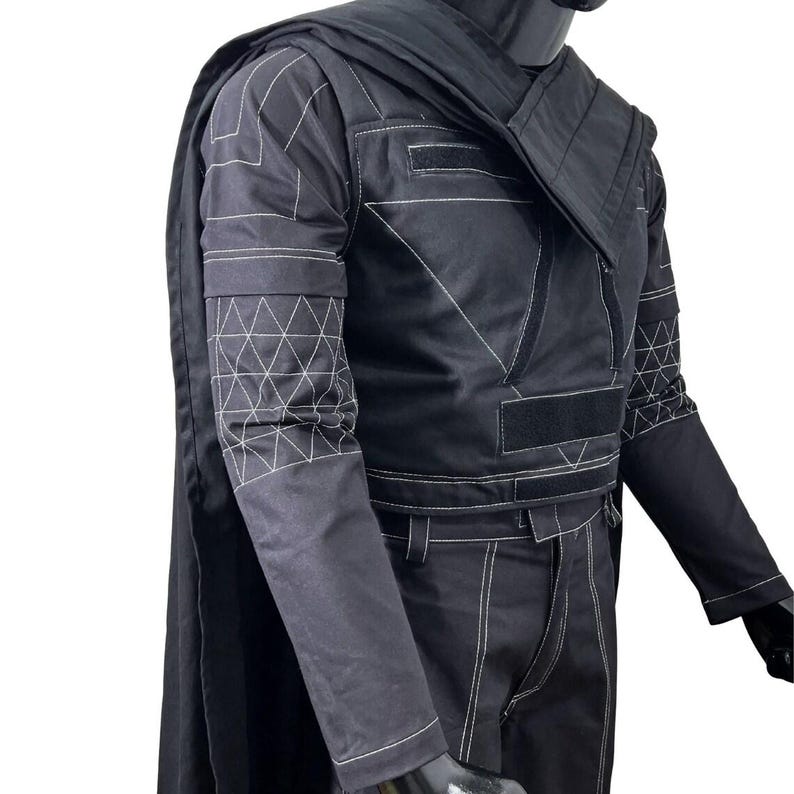 May include: A black long-sleeved jacket and matching trousers set featuring white contrast stitching. The jacket includes a structured collar, a cape attached at the shoulders, and geometric triangle patterns on the sleeves. Black velcro strips are visible on the chest area.