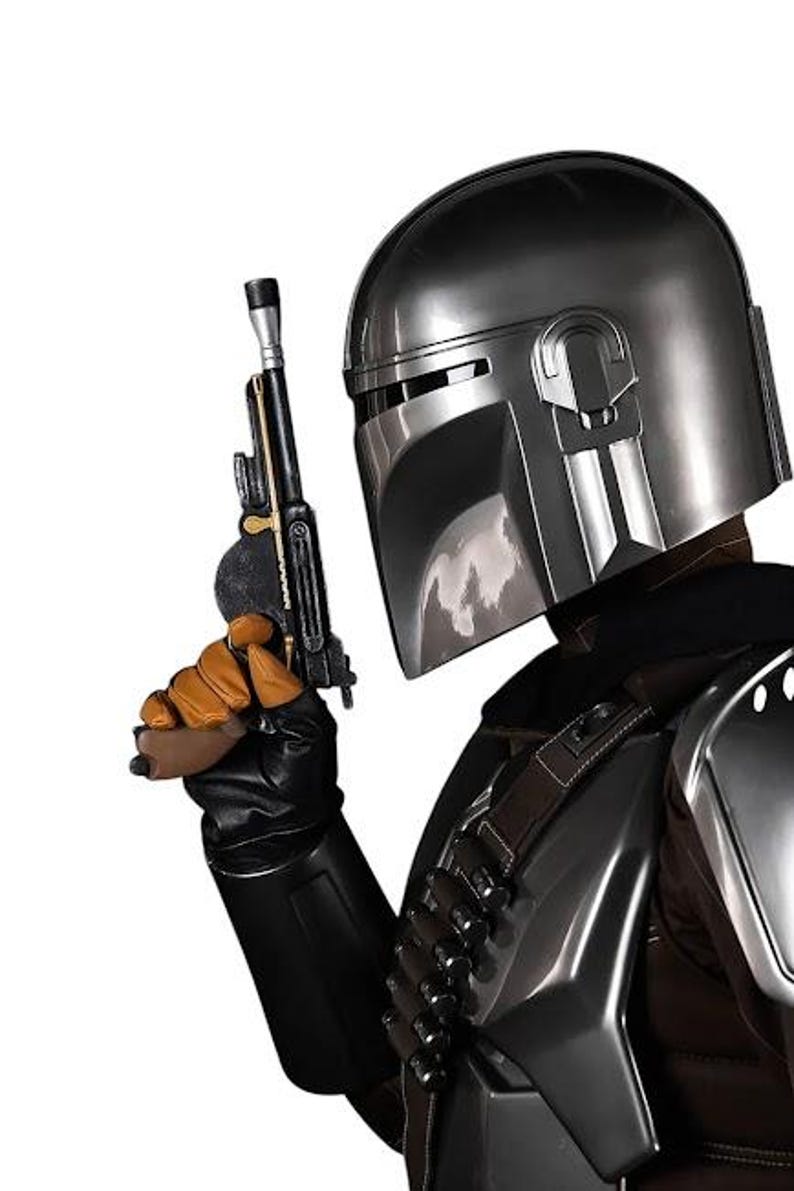 May include: A metallic silver helmet with a T-shaped visor, worn with dark armor featuring a bandolier of small cylinders. A black and gold blaster pistol is held in a gloved hand, with the armor plates reflecting light against a white background.