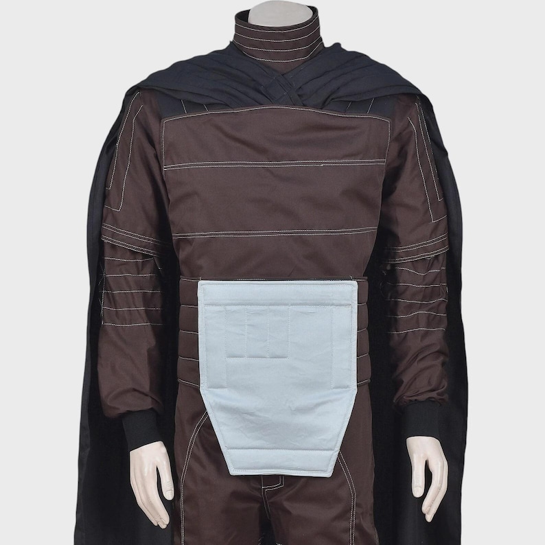 May include: A dark brown long-sleeved tunic with a high collar and white topstitching, featuring a light gray rectangular padded panel at the waist. A black cape is draped over the shoulders, and the garment is displayed on a mannequin torso.