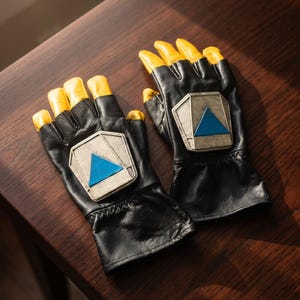 May include: A pair of black fingerless gloves featuring yellow fingertips and a metallic silver-toned pentagonal plate on the back of each hand. Each plate is centered with a bright blue triangle, and the gloves have a gathered wrist design.