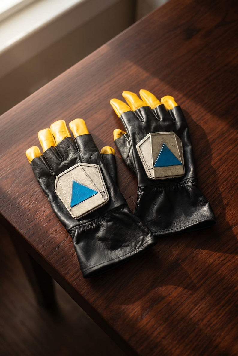 May include: A pair of black fingerless gloves featuring yellow fingertips and a metallic silver-toned pentagonal plate on the back of each hand. Each plate is centered with a bright blue triangle, and the gloves have a gathered wrist design.