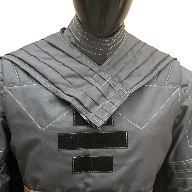 Puede incluir: Una t&uacute;nica gris de manga larga con cuello alto con costuras blancas horizontales, un dise&ntilde;o de hombros plisados en capas y tres tiras rectangulares negras de cierre de gancho y bucle colocadas verticalmente en el centro del pecho.