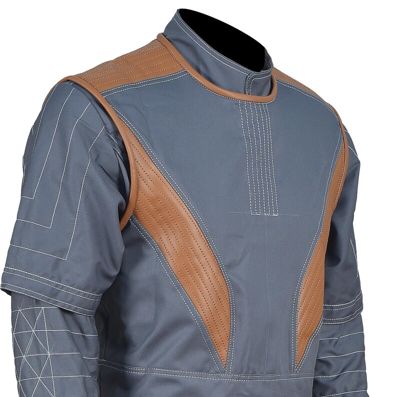 May include: A grey short-sleeved shirt featuring tan leather-like panels on the chest and shoulders. The garment includes a high collar, white topstitching details, and geometric white line patterns on the sleeves and chest area.