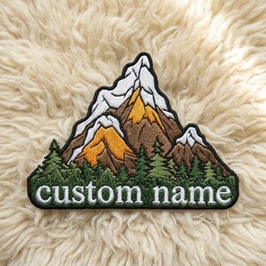 May include: Embroidered patch featuring a mountain range with snow-capped peaks, brown and yellow slopes, and green pine trees. The patch has the text "custom name" in white letters on a green border.