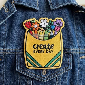 May include: An embroidered patch featuring a yellow crayon box filled with colorful flowers and crayons. The text 'create EVERY DAY' is stitched in black in the center. The patch is attached to a dark blue denim jacket with visible stitching.