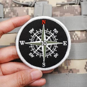 May include: A circular embroidered patch featuring a black background with a white compass rose design. The compass includes cardinal directions N, S, E, W, NE, NW, SE, and SW in white, with a red N. The center displays the number 10 in black.