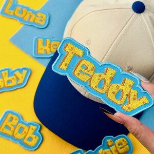 May include: A cream-colored baseball cap with a navy blue brim and a blue button on top. The cap is shown with several embroidered name patches in yellow and blue, including "Teddy", "Luna", "He", "Bobby", and "Bob". The background is yellow and blue.