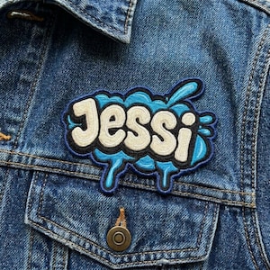May include: An embroidered patch featuring the name Jessi in cream-colored, bubble-style lettering with a thick black outline. The text is set against a bright blue, liquid-like splash design with dark blue borders, attached to a blue denim jacket.