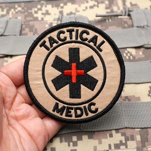 May include: A circular embroidered patch with a tan background and a black border. It features the black text TACTICAL MEDIC arched around a central black Star of Life symbol with a small, bright red cross at its center, all stitched in thread.