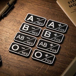 May include: A set of eight rectangular black patches with white embroidered text representing blood types. Each patch features a large letter A, B, AB, or O, paired with a plus or minus sign and the text POS or NEG, indicating positive or negative.